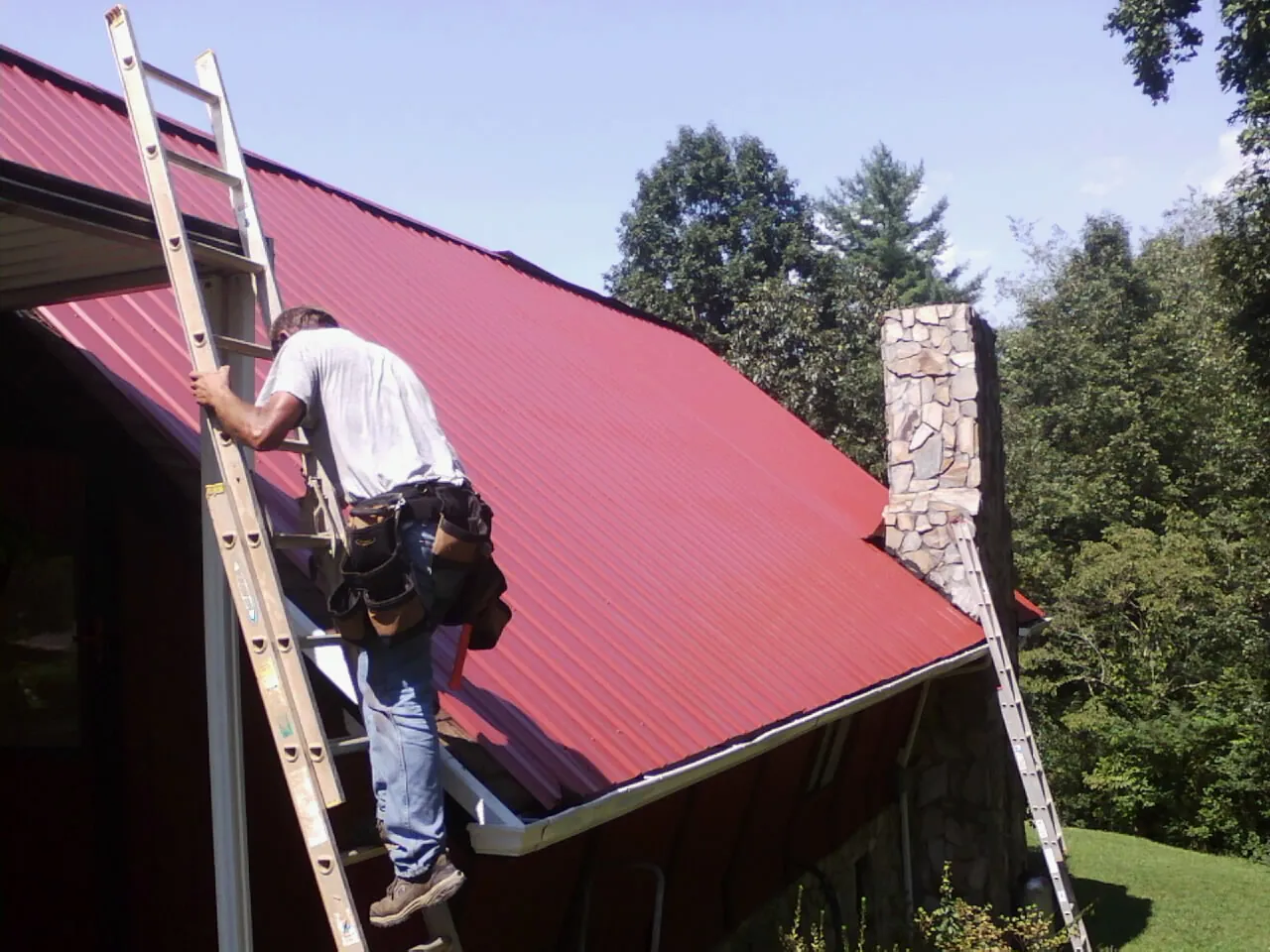 Roofer on a ladder beside a red metal roof and stone chimney — Soffit & Fascia Repair by Roofing Brothers in Rosanky